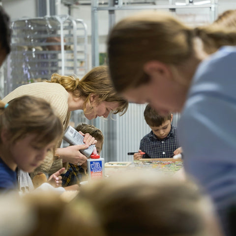 Art Instructor gathered around a table with adults and children making a collaborative mosaic artwork.